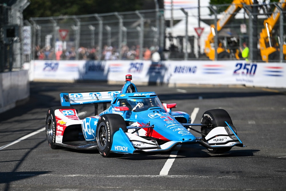 An IndyCar racing vehicle in a blue, red, and white livery is captured mid-race on a street circuit. The car prominently displays sponsor logos including Hitachi, PPG, and McLaughlin branding, with Firestone tires visible. The background shows safety fencing, spectators, race barriers with '250' branding, and construction equipment.