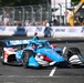 An IndyCar racing vehicle in a blue, red, and white livery is captured mid-race on a street circuit. The car prominently displays sponsor logos including Hitachi, PPG, and McLaughlin branding, with Firestone tires visible. The background shows safety fencing, spectators, race barriers with '250' branding, and construction equipment.