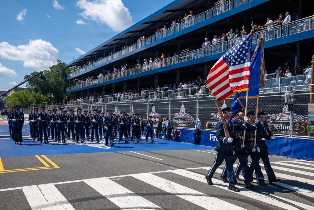 A military honor guard in U.S. Air Force dress blues marches across a racing circuit start/finish line during a pre-race ceremony. The event is identified as the 'Freedom 250 Grand Prix,' with signage visible along the pit lane barrier. Multi-tiered grandstands packed with spectators line the track in the background.