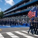 A military honor guard in U.S. Air Force dress blues marches across a racing circuit start/finish line during a pre-race ceremony. The event is identified as the 'Freedom 250 Grand Prix,' with signage visible along the pit lane barrier. Multi-tiered grandstands packed with spectators line the track in the background.