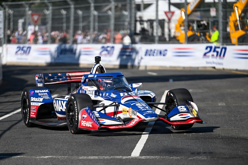 An IndyCar racing car with a patriotic American flag-themed livery sponsored by RE/MAX is competing on a street circuit. The car features red, white, and blue colors with stars and stripes design elements. Spectators and safety barriers are visible in the background along with Boeing and '250' event branding.