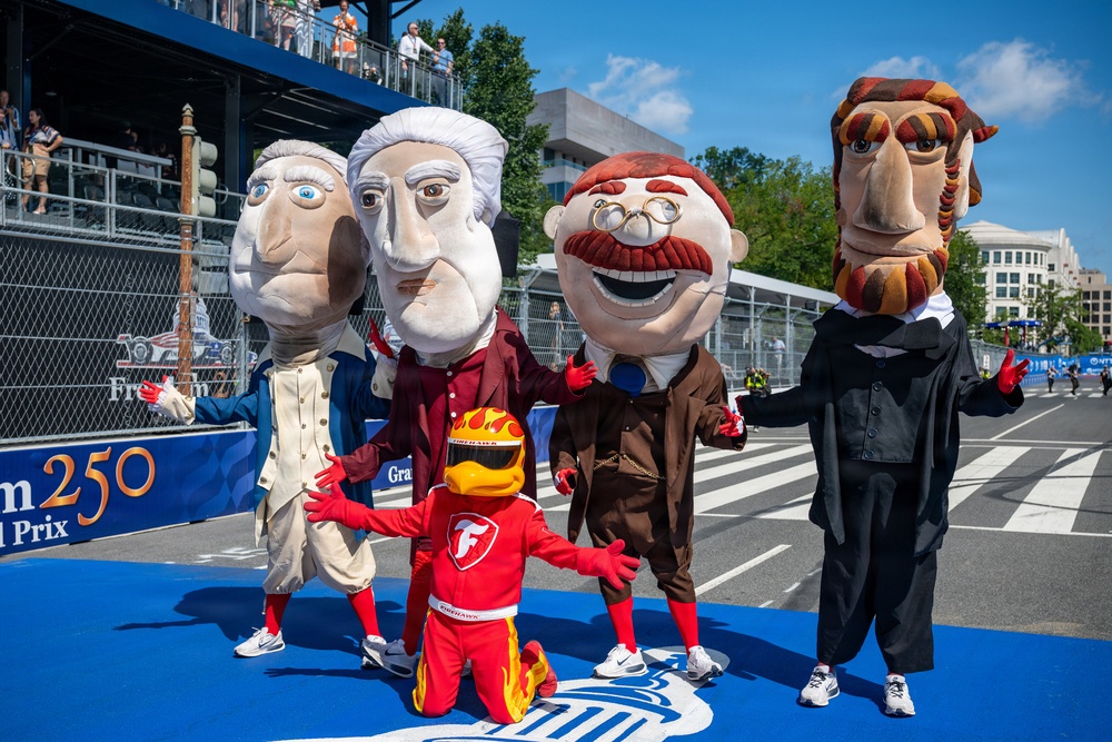 A group of oversized mascot costumes depicting historical U.S. presidents are posed together on a racing circuit track. A smaller racing mascot in a red suit and flame-decorated helmet kneels in front of them. The setting appears to be a motorsport event, with grandstands, barriers, and signage reading '250 Grand Prix' visible in the background.