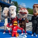 A group of oversized mascot costumes depicting historical U.S. presidents are posed together on a racing circuit track. A smaller racing mascot in a red suit and flame-decorated helmet kneels in front of them. The setting appears to be a motorsport event, with grandstands, barriers, and signage reading '250 Grand Prix' visible in the background.