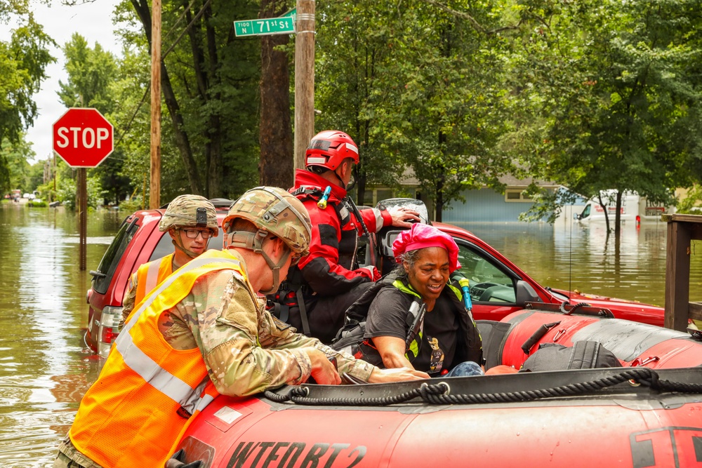 A flood rescue operation is underway at the intersection of Tioo and N 71st Street, where military personnel and emergency responders are evacuating a civilian woman from a red inflatable rescue boat. The surrounding neighborhood is heavily flooded, with vehicles partially submerged in brown floodwater. Multiple rescue personnel in military camouflage, high-visibility vests, and red rescue gear are actively assisting the evacuee.