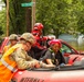 A flood rescue operation is underway at the intersection of Tioo and N 71st Street, where military personnel and emergency responders are evacuating a civilian woman from a red inflatable rescue boat. The surrounding neighborhood is heavily flooded, with vehicles partially submerged in brown floodwater. Multiple rescue personnel in military camouflage, high-visibility vests, and red rescue gear are actively assisting the evacuee.