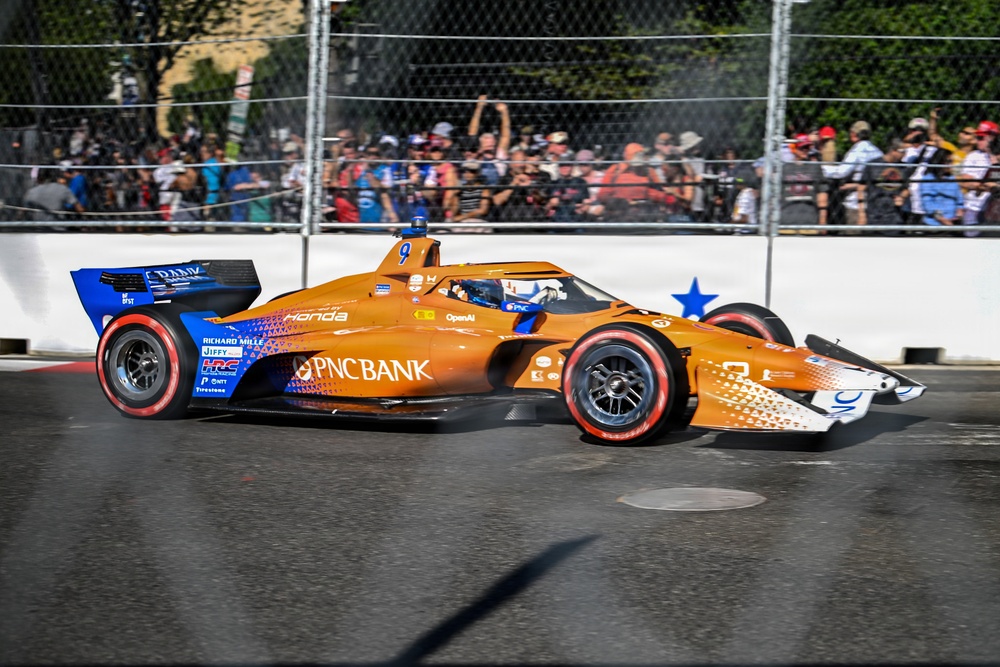 An IndyCar racing car with an orange and blue livery sponsored by PNC Bank is captured mid-race on a street circuit. The car, powered by Honda and bearing the number 9, is navigating a corner with spectators visible behind safety fencing in the background. The image is sharp and well-lit, suggesting daytime racing conditions.