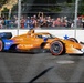 An IndyCar racing car with an orange and blue livery sponsored by PNC Bank is captured mid-race on a street circuit. The car, powered by Honda and bearing the number 9, is navigating a corner with spectators visible behind safety fencing in the background. The image is sharp and well-lit, suggesting daytime racing conditions.