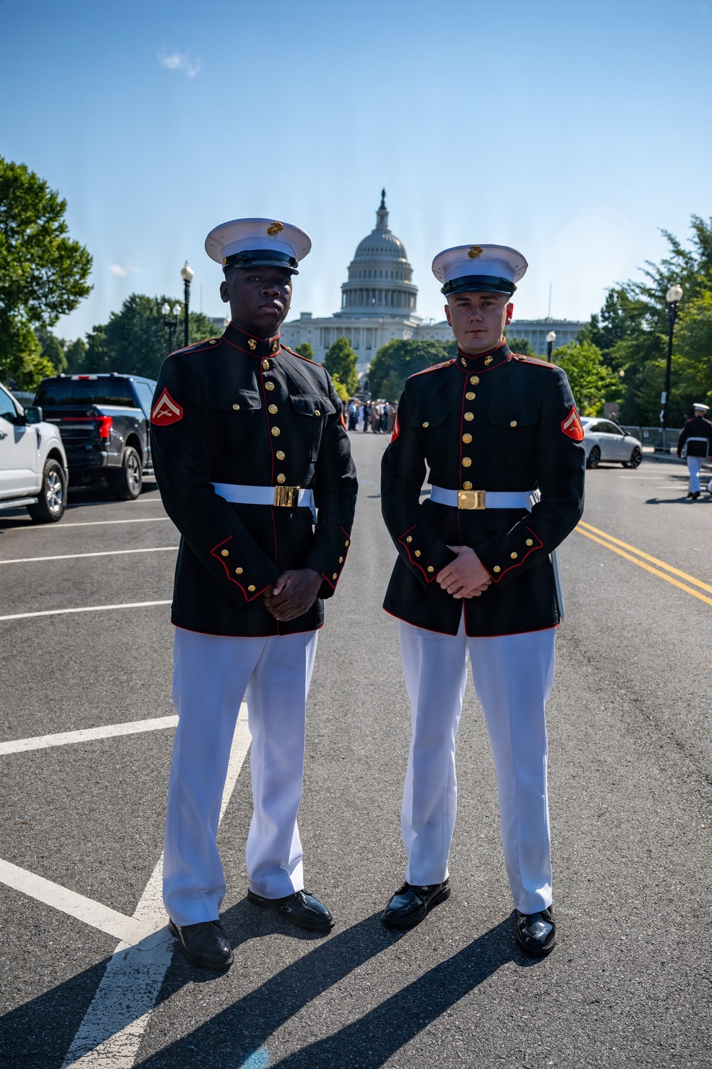 Two U.S. Marines in dress blue uniform stand at attention on a street directly in front of the United States Capitol building in Washington, D.C. Both wear the distinctive Marine Corps dress blues with white covers, white trousers, white belts, and gold buttons. The image appears to be taken during a formal ceremonial event given the road closure and additional personnel visible in the background.