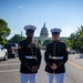 Two U.S. Marines in dress blue uniform stand at attention on a street directly in front of the United States Capitol building in Washington, D.C. Both wear the distinctive Marine Corps dress blues with white covers, white trousers, white belts, and gold buttons. The image appears to be taken during a formal ceremonial event given the road closure and additional personnel visible in the background.