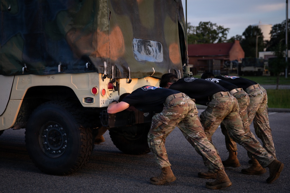 Multiple military personnel in camouflage pants and black unit t-shirts are pushing a military HMMWV (Humvee) from the rear on a paved surface. The activity appears to be a physical training exercise or vehicle recovery drill conducted at dusk or dawn. The vehicle bears the marking 'TP 42' and has a camouflage soft-top cover.