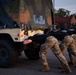 Multiple military personnel in camouflage pants and black unit t-shirts are pushing a military HMMWV (Humvee) from the rear on a paved surface. The activity appears to be a physical training exercise or vehicle recovery drill conducted at dusk or dawn. The vehicle bears the marking 'TP 42' and has a camouflage soft-top cover.