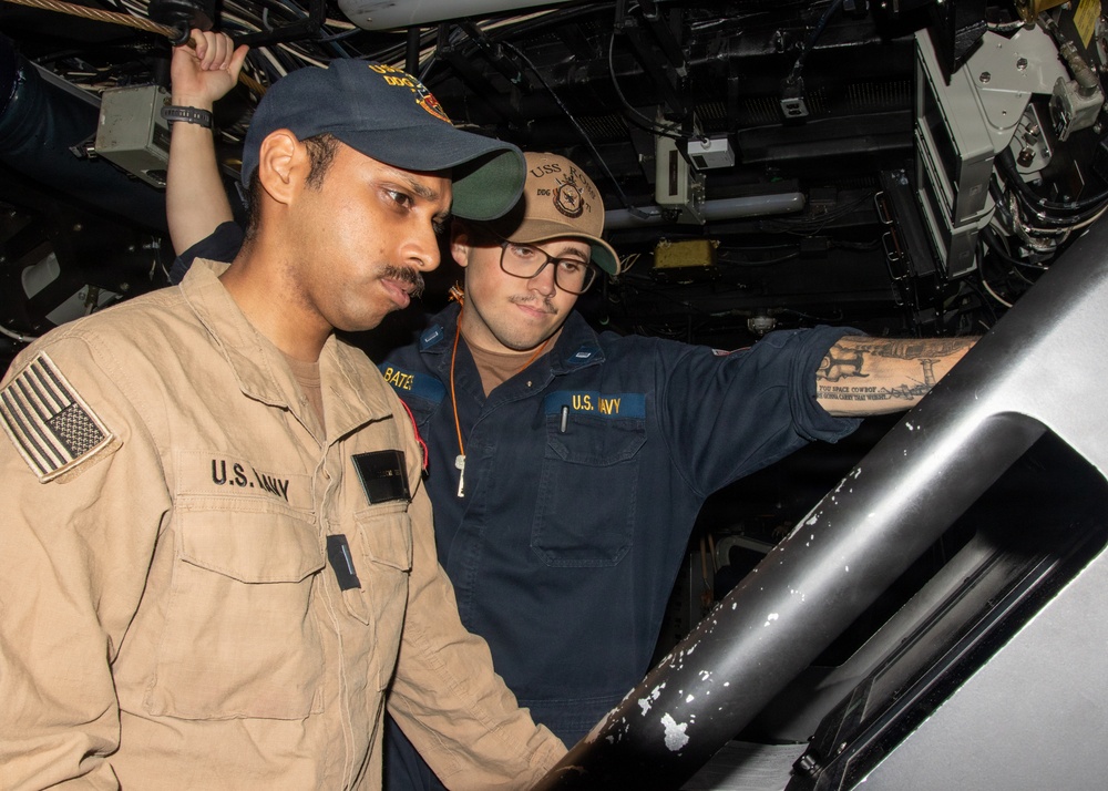 Two U.S. Navy sailors are working together in what appears to be a confined mechanical or equipment space aboard a naval vessel. The sailor on the left is wearing a tan/khaki uniform with a U.S. Navy patch and an American flag patch, while the sailor on the right wears a navy blue uniform labeled 'U.S. NAVY' with the name 'BATES' visible. Both are wearing USS-designated ball caps and appear to be examining or operating equipment.