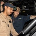 Two U.S. Navy sailors are working together in what appears to be a confined mechanical or equipment space aboard a naval vessel. The sailor on the left is wearing a tan/khaki uniform with a U.S. Navy patch and an American flag patch, while the sailor on the right wears a navy blue uniform labeled 'U.S. NAVY' with the name 'BATES' visible. Both are wearing USS-designated ball caps and appear to be examining or operating equipment.