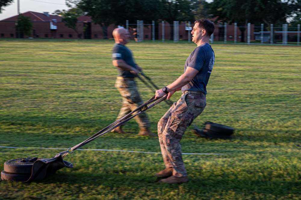 Two military personnel are conducting a physical training exercise on a grass field, pulling weighted sleds using straps. The foreground subject wears a blue t-shirt and camouflage pants, while the background subject wears a dark shirt and camouflage pants. The image is captured with motion blur on the background figure, suggesting dynamic movement during early morning or evening light.