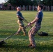 Two military personnel are conducting a physical training exercise on a grass field, pulling weighted sleds using straps. The foreground subject wears a blue t-shirt and camouflage pants, while the background subject wears a dark shirt and camouflage pants. The image is captured with motion blur on the background figure, suggesting dynamic movement during early morning or evening light.