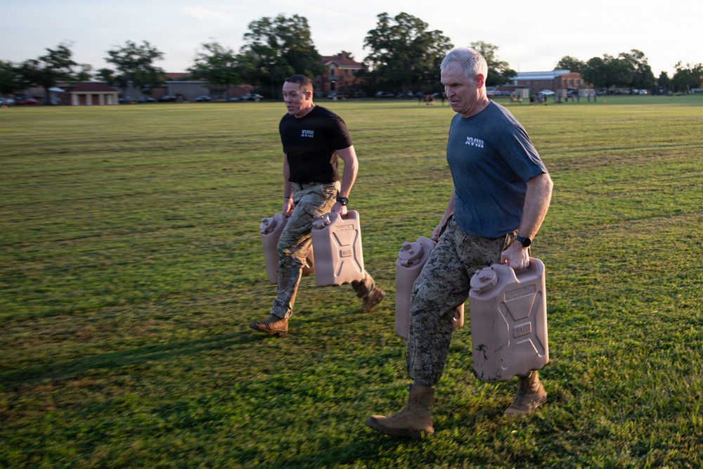 Two military personnel in physical training attire are running across a grass field while carrying tan military-issue water jerry cans. Both individuals wear matching unit t-shirts with 'XVIII' visible on the chest, along with military camouflage trousers and combat boots. The setting appears to be a military installation based on the brick buildings visible in the background.