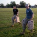 Two military personnel in physical training attire are running across a grass field while carrying tan military-issue water jerry cans. Both individuals wear matching unit t-shirts with 'XVIII' visible on the chest, along with military camouflage trousers and combat boots. The setting appears to be a military installation based on the brick buildings visible in the background.