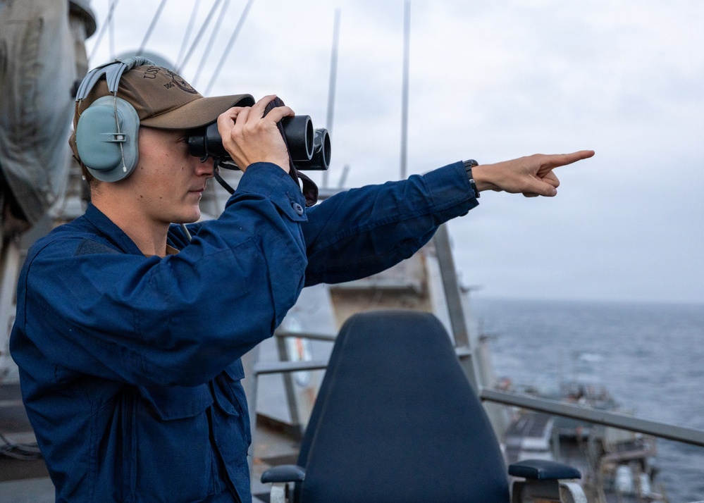 A naval personnel member stands on the bridge wing of a military vessel, looking through binoculars while simultaneously pointing in a specific direction. The individual is wearing standard Navy working uniform (blue coveralls), hearing protection headset, and a cap with partial 'US' text visible. The scene appears to be an active lookout or contact-reporting duty at sea under overcast conditions.