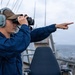 A naval personnel member stands on the bridge wing of a military vessel, looking through binoculars while simultaneously pointing in a specific direction. The individual is wearing standard Navy working uniform (blue coveralls), hearing protection headset, and a cap with partial 'US' text visible. The scene appears to be an active lookout or contact-reporting duty at sea under overcast conditions.