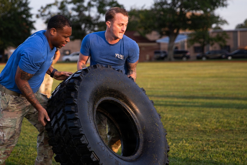 Two individuals in matching blue t-shirts and camouflage pants are performing a tire flip exercise on a grass field. The exercise appears to be part of a military or fitness training session, with both participants working together to flip a large, heavy off-road tire. The setting appears to be a military base or training facility, visible in the background.