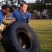 Two individuals in matching blue t-shirts and camouflage pants are performing a tire flip exercise on a grass field. The exercise appears to be part of a military or fitness training session, with both participants working together to flip a large, heavy off-road tire. The setting appears to be a military base or training facility, visible in the background.