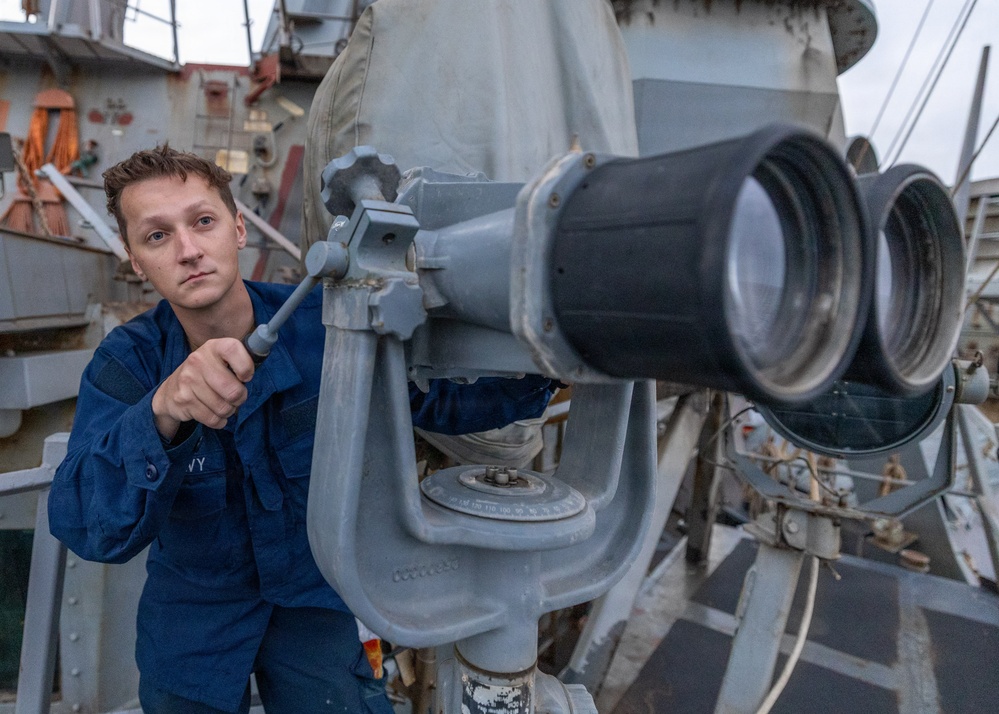 A naval service member in blue coveralls is operating or adjusting a large mounted binocular/optical observation device on the deck of a military vessel. The equipment appears to be a gyro-stabilized or pedestal-mounted big-eye binocular system commonly used for maritime surveillance. The setting is clearly aboard a warship, with gray naval superstructure visible in the background.
