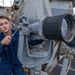 A naval service member in blue coveralls is operating or adjusting a large mounted binocular/optical observation device on the deck of a military vessel. The equipment appears to be a gyro-stabilized or pedestal-mounted big-eye binocular system commonly used for maritime surveillance. The setting is clearly aboard a warship, with gray naval superstructure visible in the background.