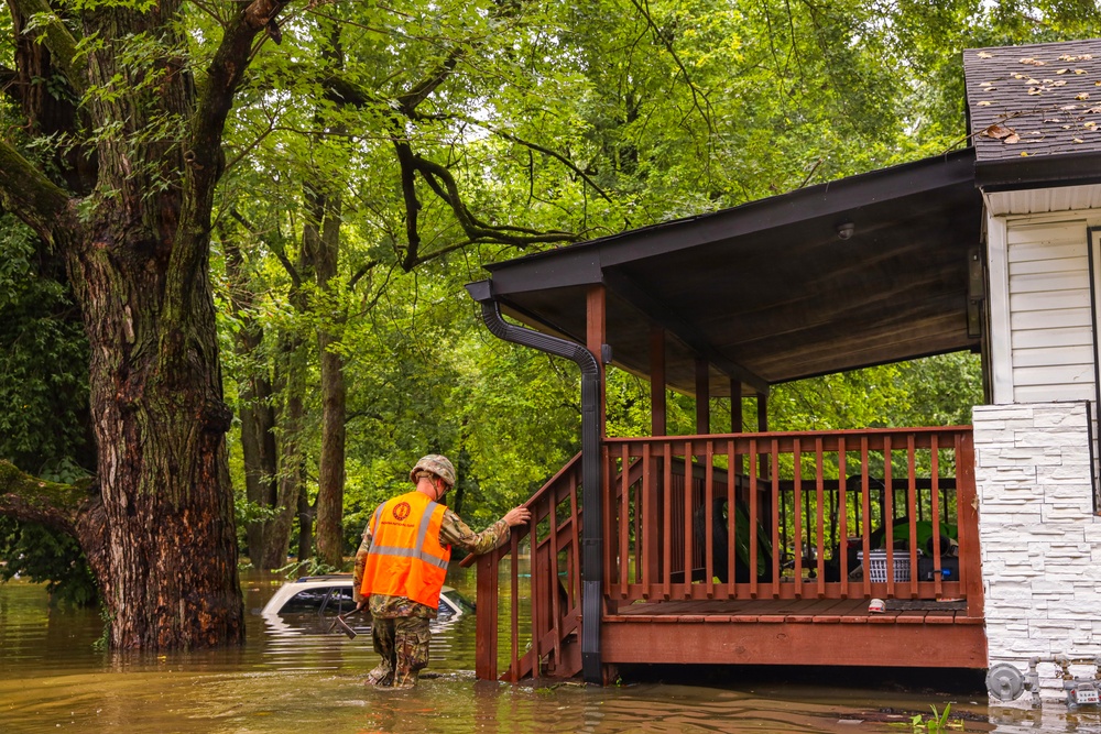 A military or emergency response personnel wearing camouflage uniform and an orange safety vest is wading through floodwaters approaching a residential home's porch. The surrounding area is heavily flooded, with a partially submerged vehicle visible in the background. The scene depicts an active flood disaster response operation in a wooded residential neighborhood.