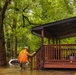 A military or emergency response personnel wearing camouflage uniform and an orange safety vest is wading through floodwaters approaching a residential home's porch. The surrounding area is heavily flooded, with a partially submerged vehicle visible in the background. The scene depicts an active flood disaster response operation in a wooded residential neighborhood.