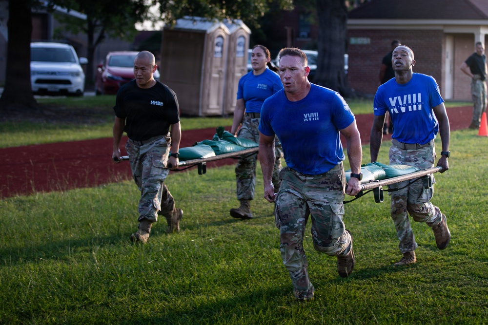 Military personnel in camouflage uniforms and blue 'XVIII' t-shirts are conducting a litter/stretcher carry exercise on a grass field. A team of four soldiers is carrying a loaded stretcher with what appears to be sandbags or weighted equipment. The activity appears to be part of a physical fitness or combat readiness training event.