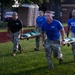 Military personnel in camouflage uniforms and blue 'XVIII' t-shirts are conducting a litter/stretcher carry exercise on a grass field. A team of four soldiers is carrying a loaded stretcher with what appears to be sandbags or weighted equipment. The activity appears to be part of a physical fitness or combat readiness training event.