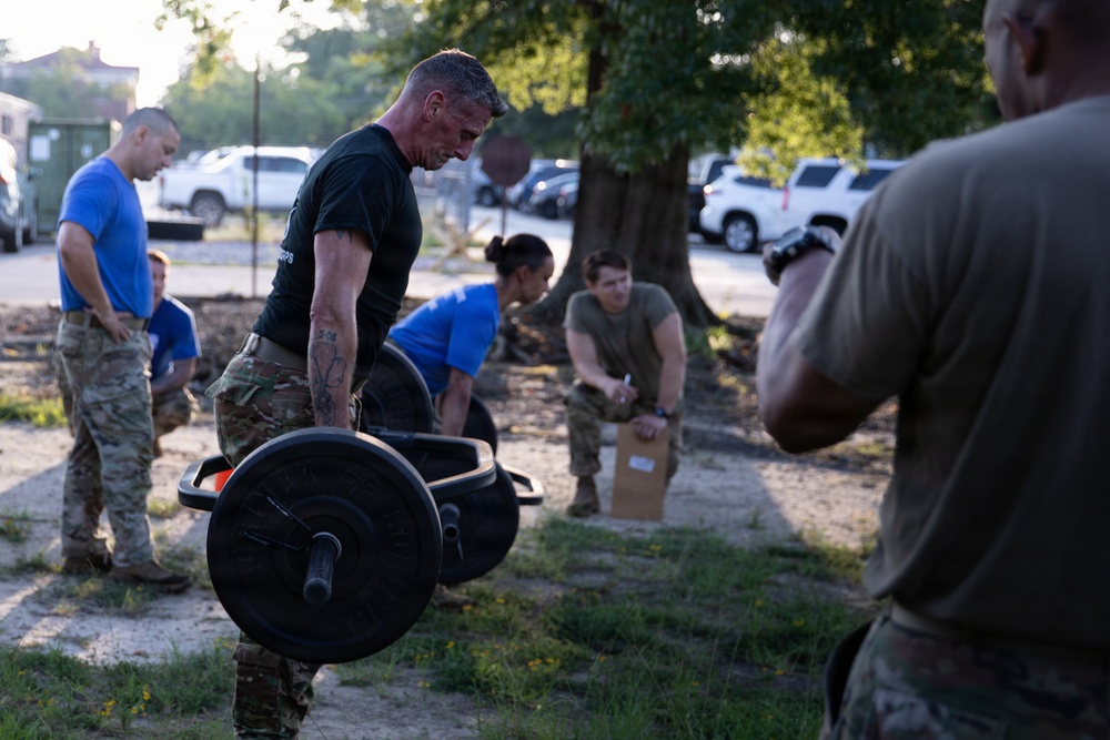 A group of military personnel are engaged in an outdoor physical training or fitness assessment exercise. A central figure in a black t-shirt and camouflage pants is performing a deadlift or carry with a loaded barbell. Other personnel in mixed uniforms — blue shirts and camouflage — are observing, recording, or participating in nearby activities.