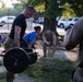 A group of military personnel are engaged in an outdoor physical training or fitness assessment exercise. A central figure in a black t-shirt and camouflage pants is performing a deadlift or carry with a loaded barbell. Other personnel in mixed uniforms — blue shirts and camouflage — are observing, recording, or participating in nearby activities.