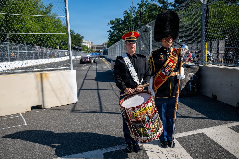 Two military band members in formal dress uniforms stand on the edge of what appears to be a street racing circuit. One member holds a decorated bass drum with what appears to be a U.S. military insignia, while the other wears a tall black bearskin hat and carries a ceremonial mace or staff. A racing car is visible in the background on the track.