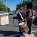 Two military band members in formal dress uniforms stand on the edge of what appears to be a street racing circuit. One member holds a decorated bass drum with what appears to be a U.S. military insignia, while the other wears a tall black bearskin hat and carries a ceremonial mace or staff. A racing car is visible in the background on the track.
