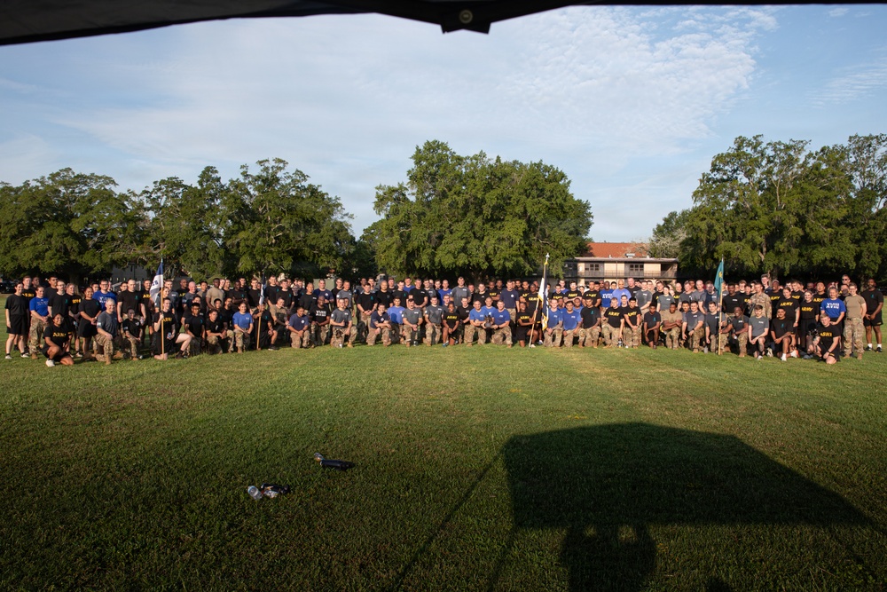 A large group photo of what appears to be U.S. Army personnel posing on a grass field, likely on a military installation. The group consists of hundreds of individuals wearing a mix of military physical training attire (black and blue 'ARMY' t-shirts) and camouflage uniforms. Several unit guidon flags are visible among the formation, suggesting this is an official unit or command group photograph.