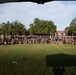 A large group photo of what appears to be U.S. Army personnel posing on a grass field, likely on a military installation. The group consists of hundreds of individuals wearing a mix of military physical training attire (black and blue 'ARMY' t-shirts) and camouflage uniforms. Several unit guidon flags are visible among the formation, suggesting this is an official unit or command group photograph.