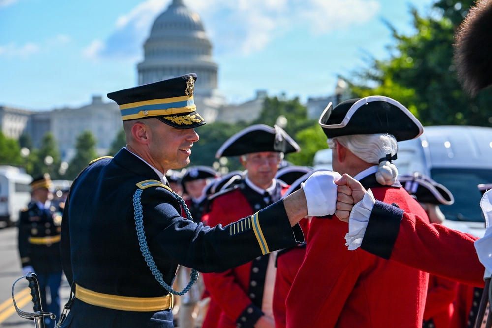 A modern U.S. Army officer in dress uniform shakes hands with a person dressed in 18th-century Continental Army/Revolutionary War period costume near the U.S. Capitol building in Washington, D.C. The scene appears to be part of a ceremonial parade or commemorative military event. Multiple participants in both modern and historical uniforms are visible in the background.