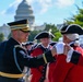 A modern U.S. Army officer in dress uniform shakes hands with a person dressed in 18th-century Continental Army/Revolutionary War period costume near the U.S. Capitol building in Washington, D.C. The scene appears to be part of a ceremonial parade or commemorative military event. Multiple participants in both modern and historical uniforms are visible in the background.