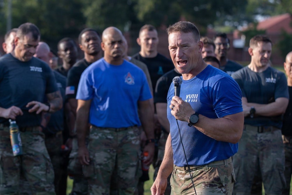 A man in a blue t-shirt and camouflage pants speaks into a microphone while addressing a large group of military personnel outdoors. The speaker appears to be delivering a motivational speech or briefing, possibly following a physical training event, as evidenced by visible perspiration. The group behind him consists of diverse military members wearing a mix of camouflage uniforms and athletic shirts.