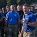 A man in a blue t-shirt and camouflage pants speaks into a microphone while addressing a large group of military personnel outdoors. The speaker appears to be delivering a motivational speech or briefing, possibly following a physical training event, as evidenced by visible perspiration. The group behind him consists of diverse military members wearing a mix of camouflage uniforms and athletic shirts.