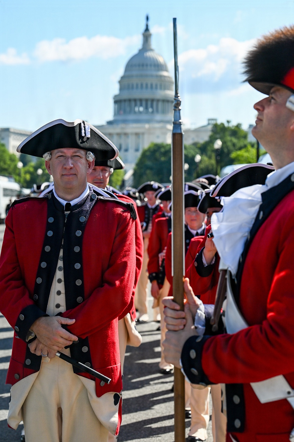 A group of individuals dressed in 18th-century American Revolutionary War-era military uniforms are assembled near the U.S. Capitol building in Washington, D.C. They are wearing red coats with black trim, tricorn hats, white wigs, and tan breeches, consistent with Continental Army or period reenactment attire. The scene appears to be a ceremonial parade or public reenactment event.
