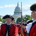 A group of individuals dressed in 18th-century American Revolutionary War-era military uniforms are assembled near the U.S. Capitol building in Washington, D.C. They are wearing red coats with black trim, tricorn hats, white wigs, and tan breeches, consistent with Continental Army or period reenactment attire. The scene appears to be a ceremonial parade or public reenactment event.