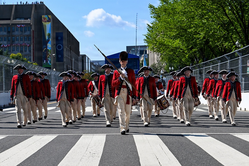 A military fife and drum corps dressed in 18th-century Revolutionary War-era uniforms marches down a city street. The performers wear red coats, cream-colored breeches, tricorn hats, and black shoes, consistent with the U.S. Army Old Guard Fife and Drum Corps. The setting appears to be a formal parade in an urban environment, likely Washington, D.C., based on the architecture and street barriers visible in the background.