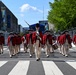 A military fife and drum corps dressed in 18th-century Revolutionary War-era uniforms marches down a city street. The performers wear red coats, cream-colored breeches, tricorn hats, and black shoes, consistent with the U.S. Army Old Guard Fife and Drum Corps. The setting appears to be a formal parade in an urban environment, likely Washington, D.C., based on the architecture and street barriers visible in the background.