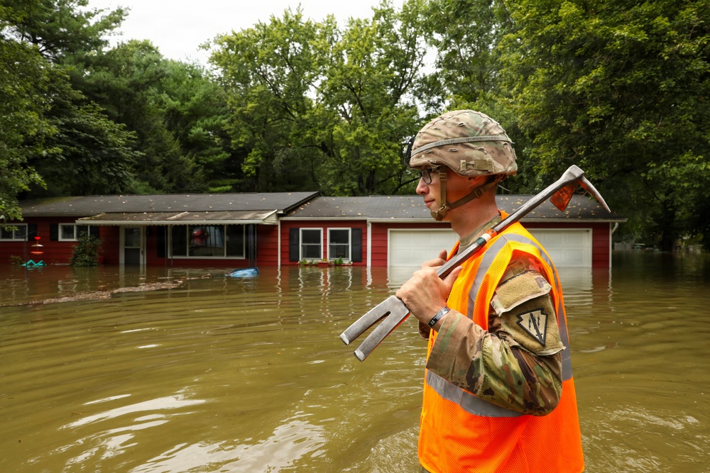 A soldier in military uniform and combat helmet, wearing a high-visibility orange safety vest, stands in floodwater in front of a severely inundated residential property. The floodwater reaches near the windows of a single-story red house, with murky brown water covering the entire yard. The soldier is carrying a pry bar or halligan tool, suggesting active search-and-rescue or damage assessment operations.
