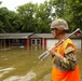 A soldier in military uniform and combat helmet, wearing a high-visibility orange safety vest, stands in floodwater in front of a severely inundated residential property. The floodwater reaches near the windows of a single-story red house, with murky brown water covering the entire yard. The soldier is carrying a pry bar or halligan tool, suggesting active search-and-rescue or damage assessment operations.