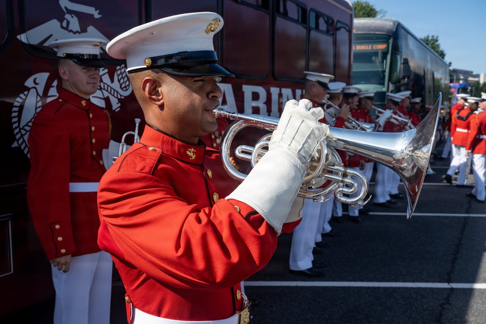 A United States Marine Corps band is assembled outdoors, with a foreground musician playing a silver bugle or flugelhorn in full dress uniform. The musicians are wearing the distinctive red and white Marine Corps dress uniform with white gloves and white covers (caps). A dark red bus with 'MARINE' lettering and 'BUGLE CORP' destination sign is visible in the background.