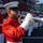 A United States Marine Corps band is assembled outdoors, with a foreground musician playing a silver bugle or flugelhorn in full dress uniform. The musicians are wearing the distinctive red and white Marine Corps dress uniform with white gloves and white covers (caps). A dark red bus with 'MARINE' lettering and 'BUGLE CORP' destination sign is visible in the background.