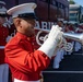 A United States Marine Corps band is assembled outdoors, with a foreground musician playing a silver bugle or flugelhorn in full dress uniform. The musicians are wearing the distinctive red and white Marine Corps dress uniform with white gloves and white covers (caps). A dark red bus with 'MARINE' lettering and 'BUGLE CORP' destination sign is visible in the background.